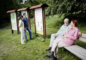Ein Mann und eine Frau sitzen auf einer Bank im Grünen, während zwei andere Menschen sich eine Informationstafel ansehen