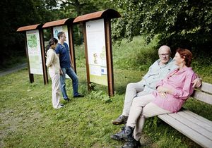 Ein Mann und eine Frau sitzen auf einer Bank im Grünen, während zwei andere Menschen sich eine Informationstafel ansehen